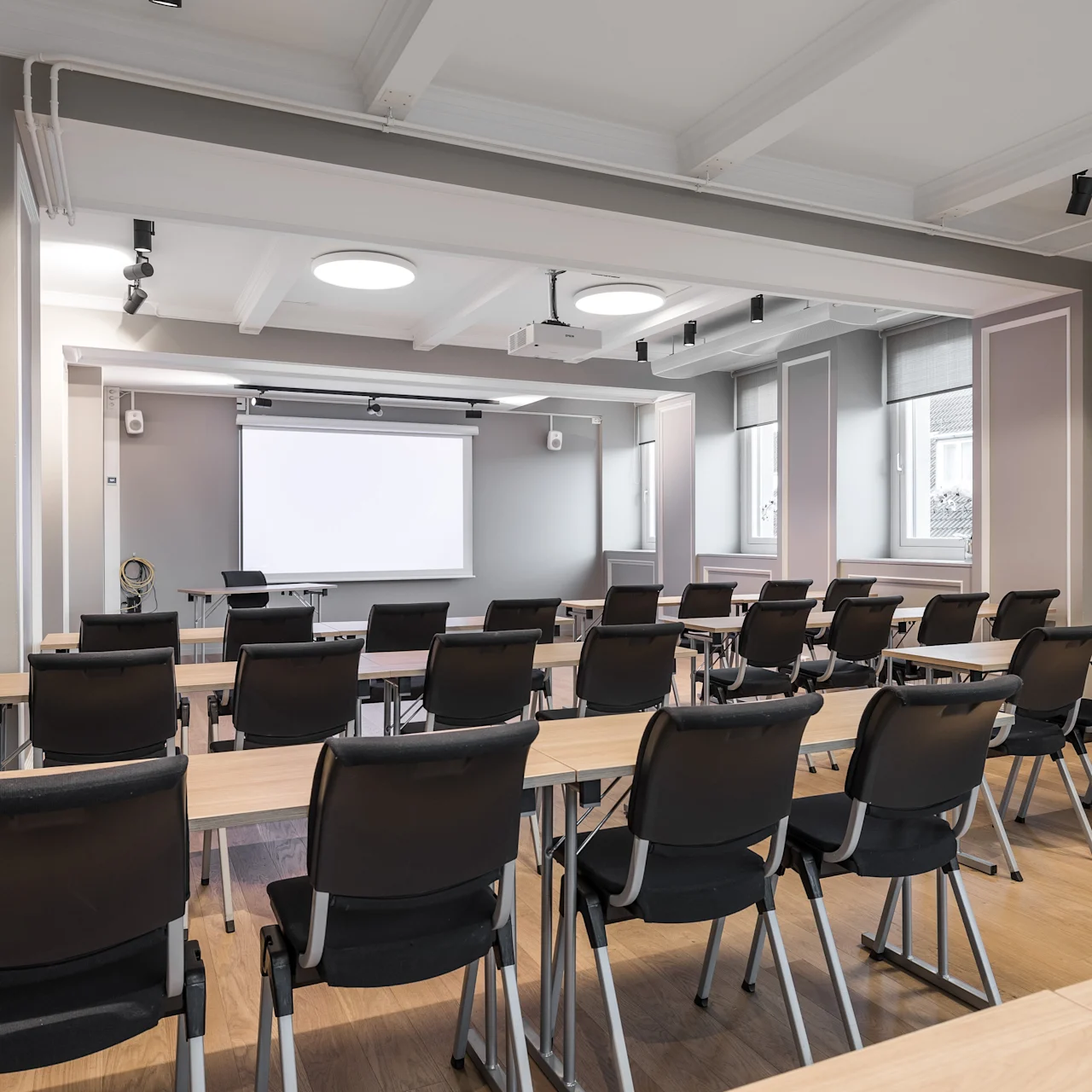 Rivit mustia tuoleja ovat vastakkain suuren valkoisen projektorikankaan kanssa valaistussa, modernissa kokoushuoneessa, jossa on puulattiat ja kattovalot.