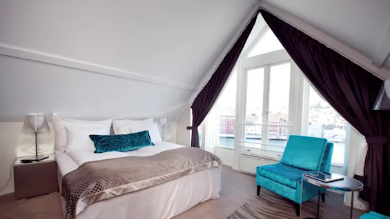 A well-lit attic bedroom with an inclined ceiling, large windows overlooking a body of water and buildings, plush bedding, and elegant furniture.