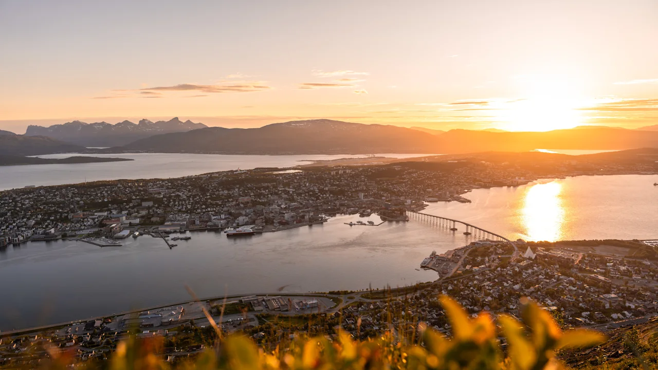 En rolig bukt reflekterer den nedgående solen; en bro forbinder en livlig bybilde med fastlandet, omgitt av fjerne fjell og opplyst av gyllent lys.