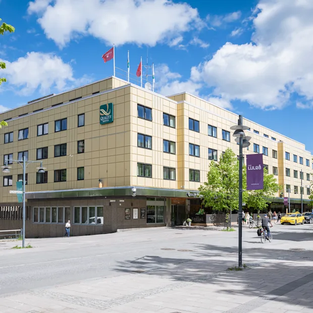 En stor hotellbyggnad med en grön logotyp under blå himmel, med människor som går och cyklar framför.
