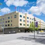 En stor hotellbyggnad med en grön logotyp under blå himmel, med människor som går och cyklar framför.