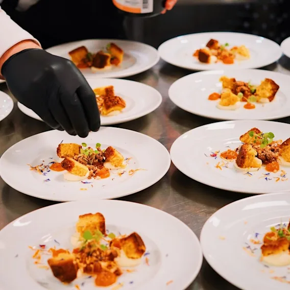 En kock med svarta handskar dekorerar flera vita tallrikar med gourmetdesserter på en rostfri köksbänk.