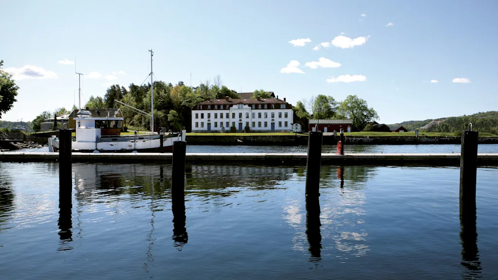 Boat moored at a waterfront, with a large white building and lush greenery in the background under a clear blue sky.