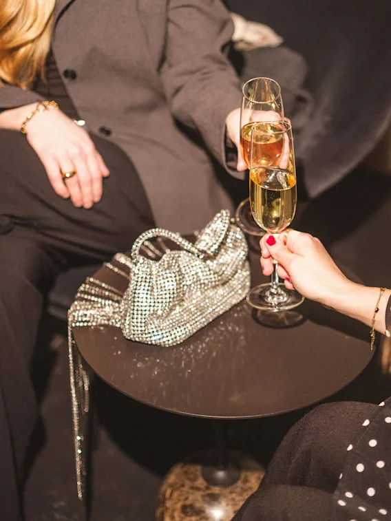 Två personer skålar med champagneglas över ett litet bord med en glittrig handväska.