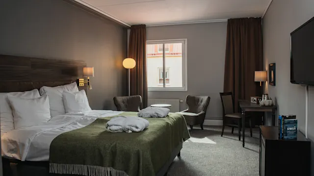 A neatly made bed with green throws and pillows sits in a hotel room with chairs, a desk, and a window with brown curtains. Soft lighting creates a cozy atmosphere.