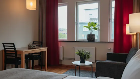 A neatly arranged room features a bed with grey bedding, a small round table with a plant, a table with chairs and a radio, under warm lighting and red curtains.