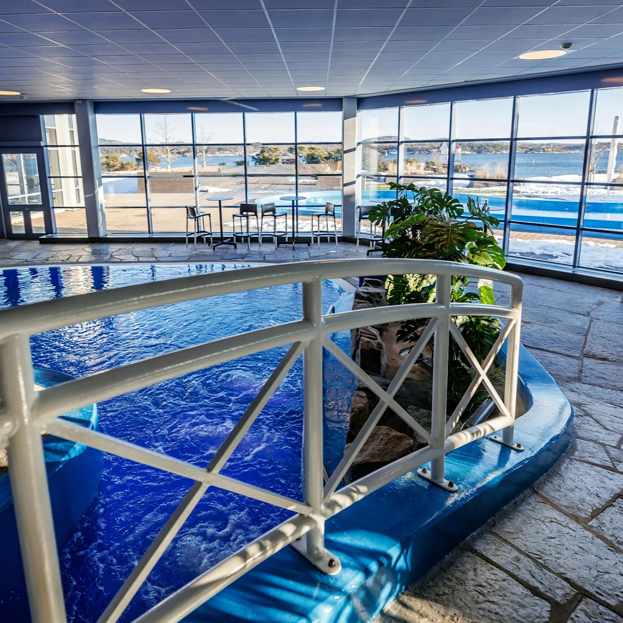 Indoor pool with clear blue water, surrounded by a white railing in a spacious room. Large windows reveal an outdoor snowy landscape with lounge chairs, creating a serene atmosphere.