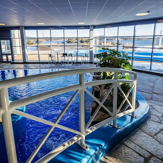 Innendørs basseng med klart blått vann, omgitt av et hvitt rekkverk i et romslig rom. Store vinduer viser et snødekket landskap utenfor med solstoler, noe som skaper en rolig atmosfære.