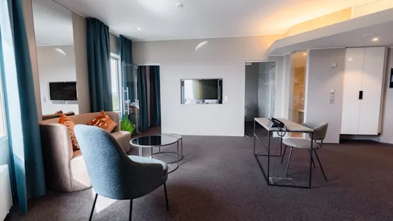 A modern room with a grey armchair and a sofa facing a wall-mounted TV, complemented by a small round table. Large windows with blue curtains illuminate the space.