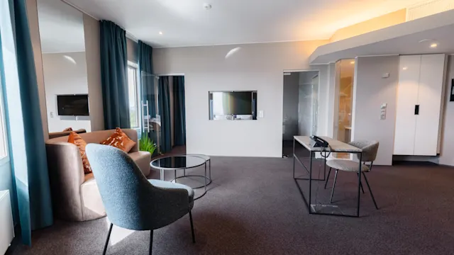 A modern room with a grey armchair and a sofa facing a wall-mounted TV, complemented by a small round table. Large windows with blue curtains illuminate the space.