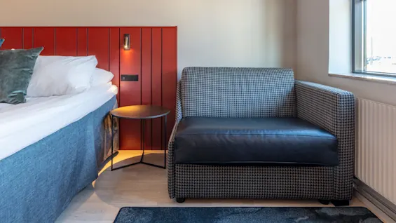 A modern sofa and bed in a neat hotel room with a red accent wall, warm lighting, and a window offering daylight.