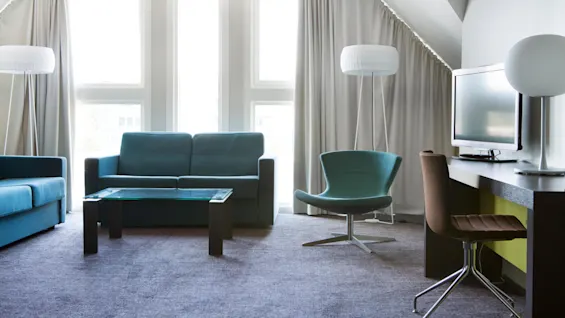 A modern living room features a blue sofa, glass coffee table, teal chair, and a desk with a TV, set against tall windows and light curtains.