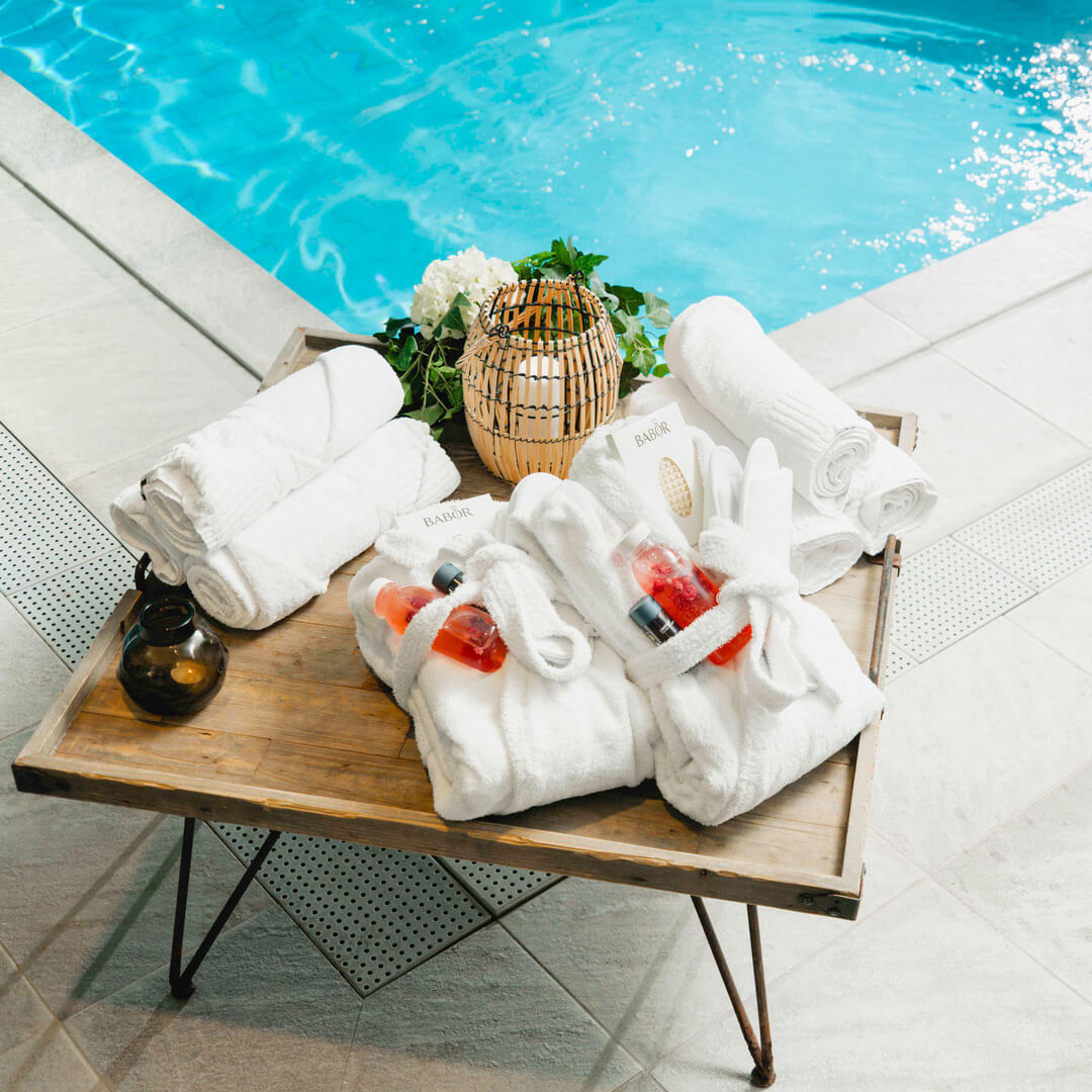 Ett träbord med spa-föremål bredvid en pool – handdukar, morgonrockar, blommor och hudvårdsprodukter.