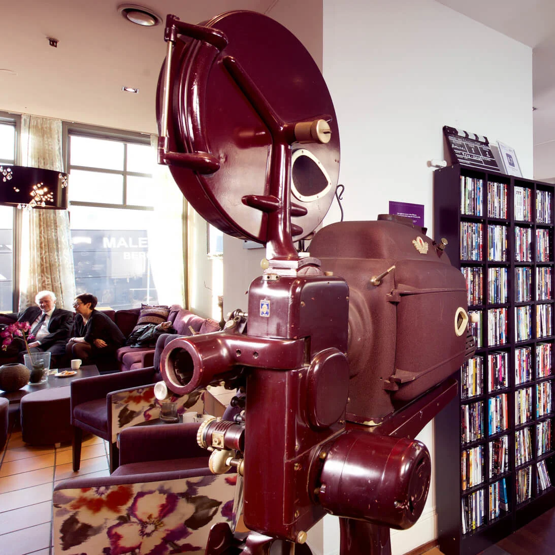 En gammeldags prosjektor står i et moderne loungeområde hvor folk sitter og prater, omgitt av DVD-er og utsikt til byen gjennom store vinduer.