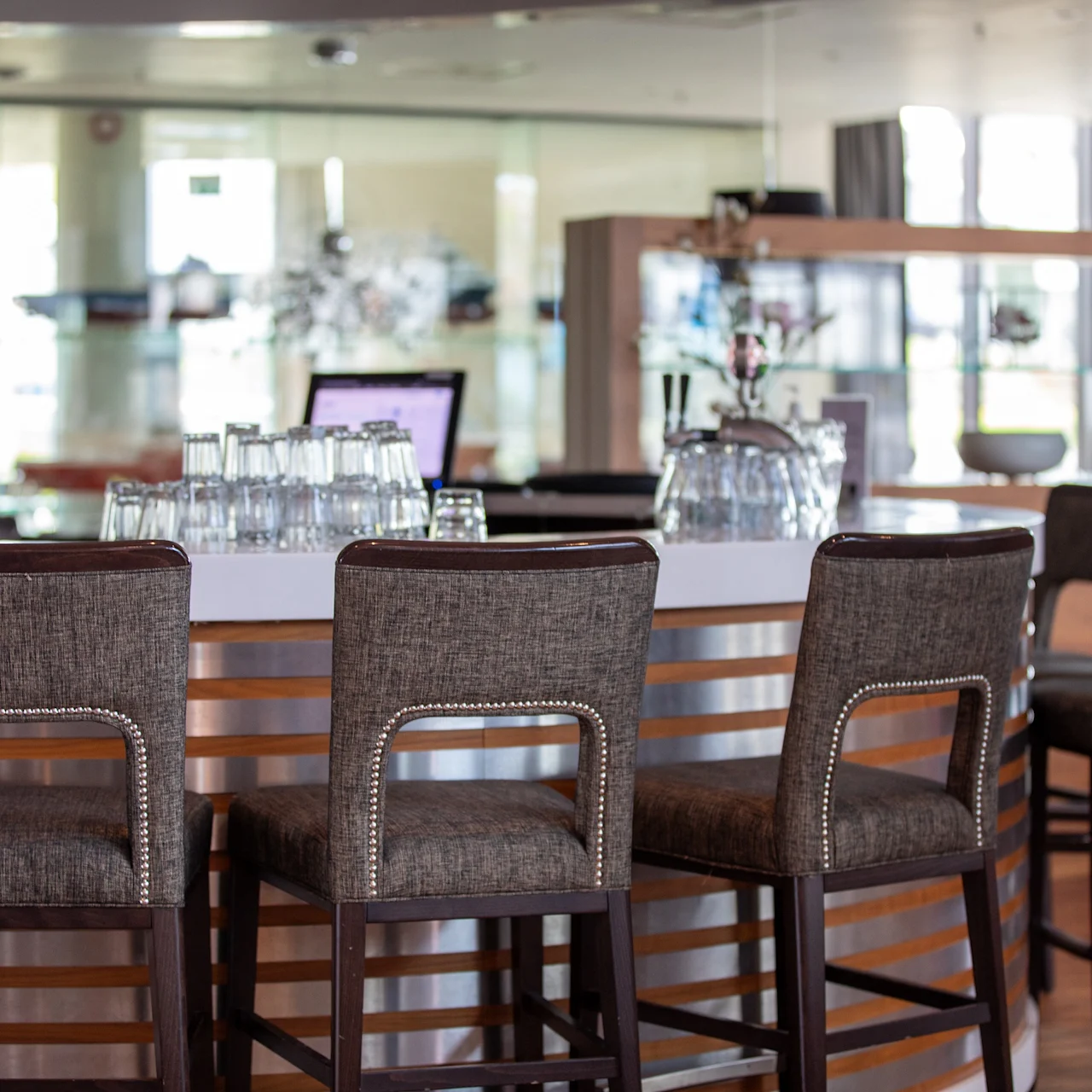 Barstoler står pent på rad langs en moderne bar, med glass plassert oppå. Den rolige, velopplyste loungen har tre detaljer og moderne dekor, og fremkaller en avslappet atmosfære.