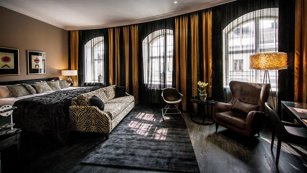Et luksuriøst hotellrom har en stor seng med dekorative puter, en sebrastrøket sofa, elegant lenestol og høy lampe. Gulv-til-tak-vinduer er dekket av mørke gardiner.