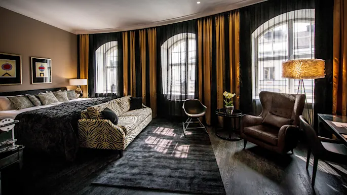 Et luksuriøst hotellrom har en stor seng med dekorative puter, en sebrastrøket sofa, elegant lenestol og høy lampe. Gulv-til-tak-vinduer er dekket av mørke gardiner.