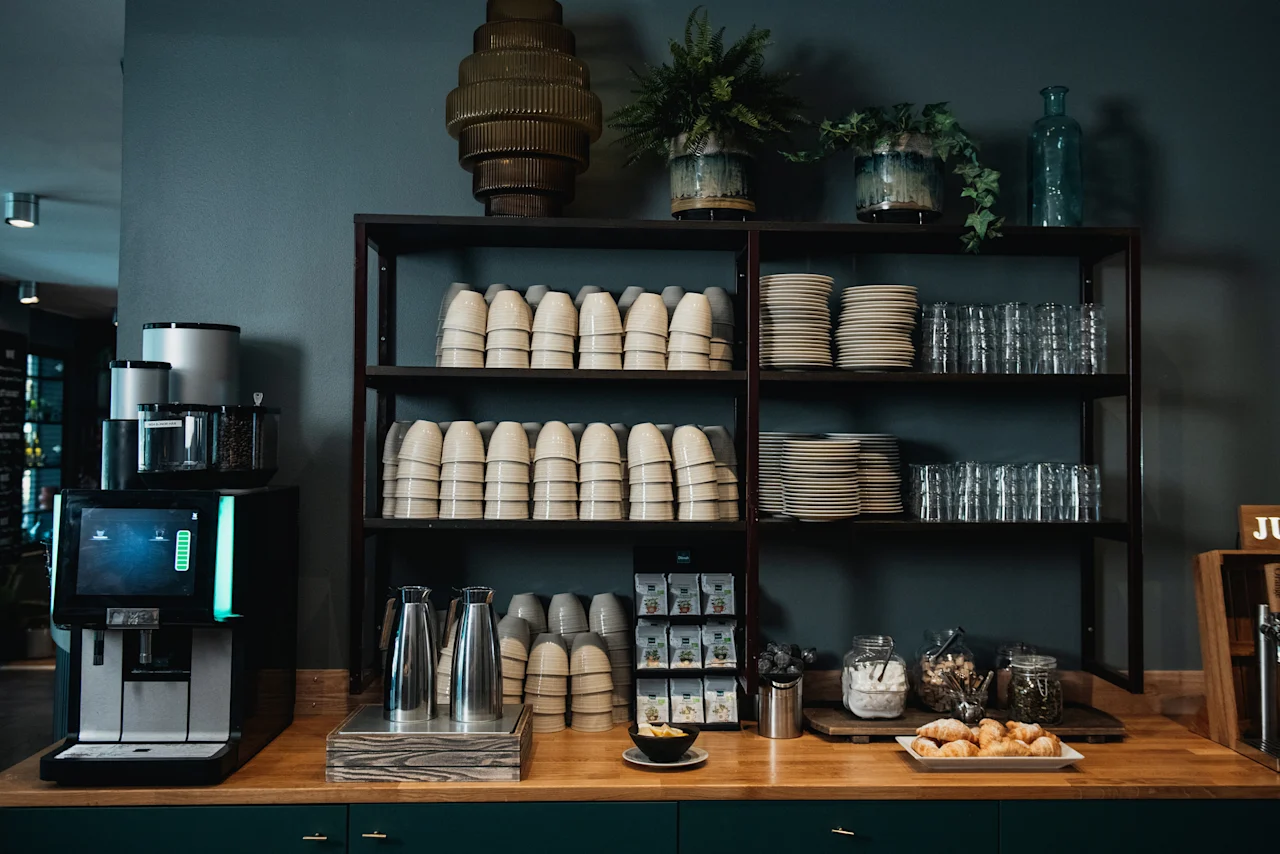 Ett välfyllt självbetjäningsområde med kaffemaskin, diverse porslin och färska bakverk på en träbänk.