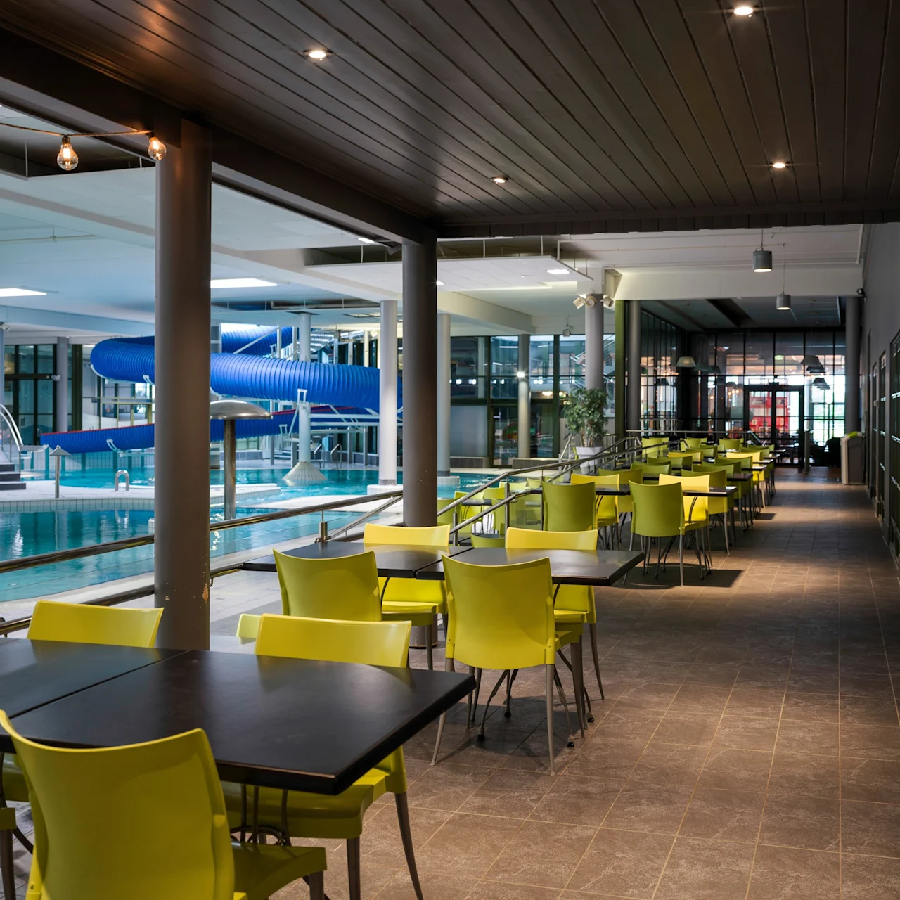 Tables and lime-green chairs line a walkway beside a swimming pool with a large blue water slide, all within an indoor leisure facility under bright ceiling lights.