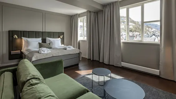 En pænt redt seng med to lamper og puder står i et moderne, rummeligt rum. En grøn sofa og runde borde er i nærheden, med store vinduer der viser en bjergrig udsigt.