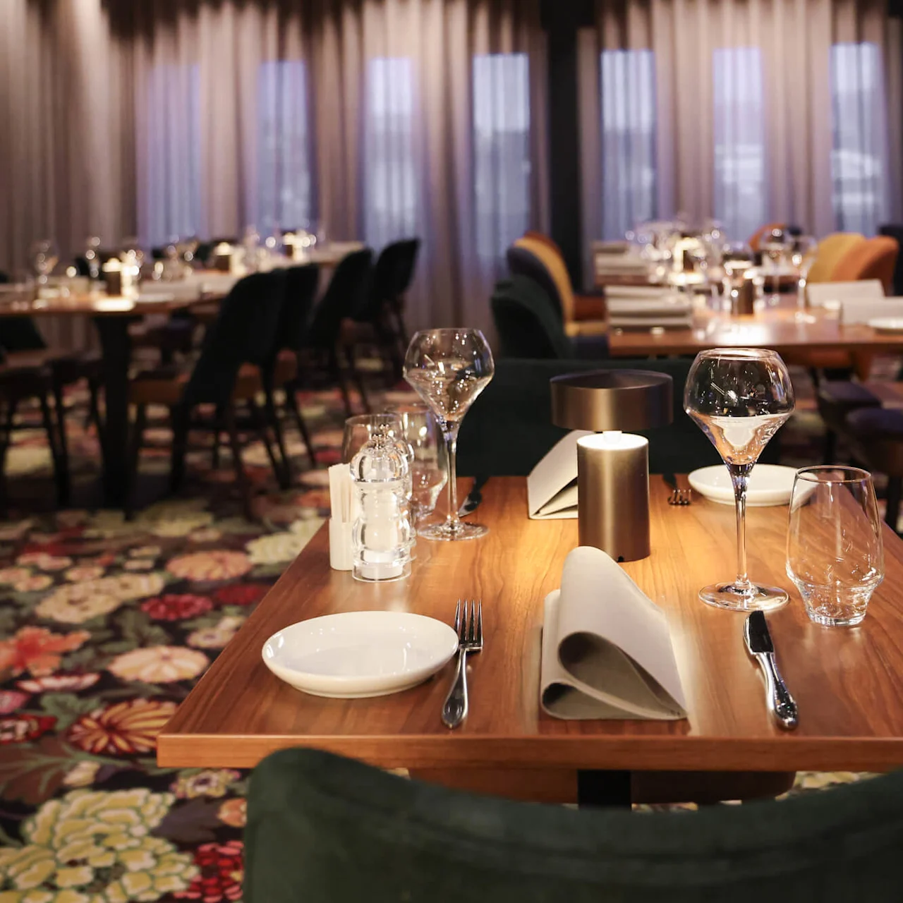 A wooden table is elegantly set with wine glasses, a candle, and cutlery on a floral carpet in a dimly lit restaurant. No text is visible.