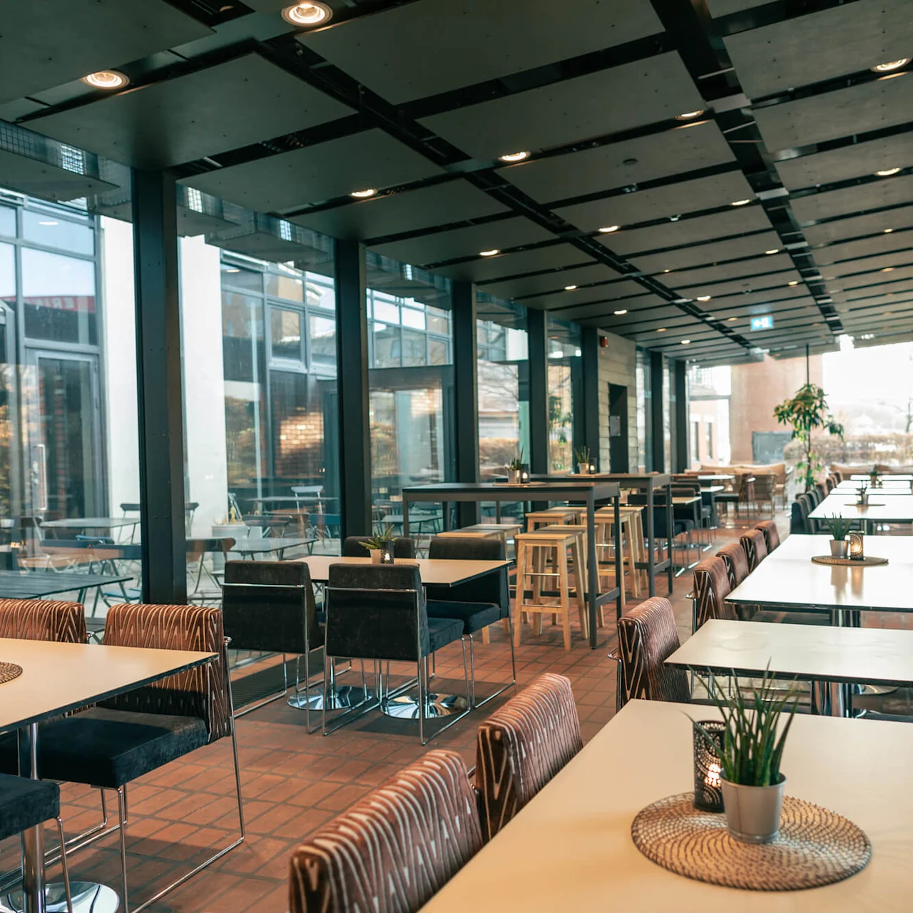Contemporary café interior features tables and chairs in a long, bright space with glass walls and brick accents; potted plants adorn tables, creating a welcoming atmosphere. No text present.