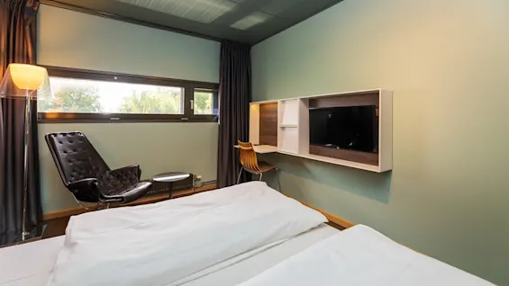 Bed lies unoccupied with white linens, beside modern black chair in minimalistic room; flat-screen TV mounted on light green wall, small window letting in natural light.