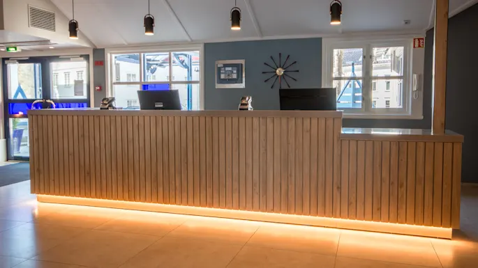 A wooden reception desk with subtle lighting stands surrounded by modern decor. Behind, windows let in daylight, and a black clock adorns the wall.
