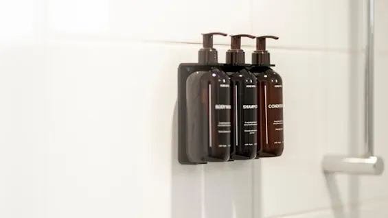 Tre mörka pumpflaskor märkta BODY WASH, SHAMPOO och CONDITIONER monterade på en vitkaklad duschvägg.