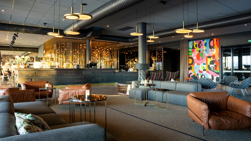 Et moderne loungeområde har brune læder- og gråstoffmøbler rundt lave borde; en elegant bar med gyldne detaljer er i baggrunden. Farverig abstrakt kunst siger "we the people."