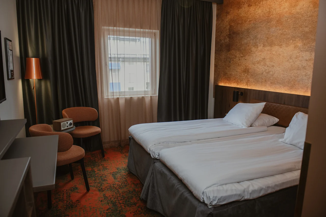 Et komfortabelt hotellrom med to pent oppredde senger, et vindu med både tynne og mørkleggende gardiner, og et lite sitteområde.