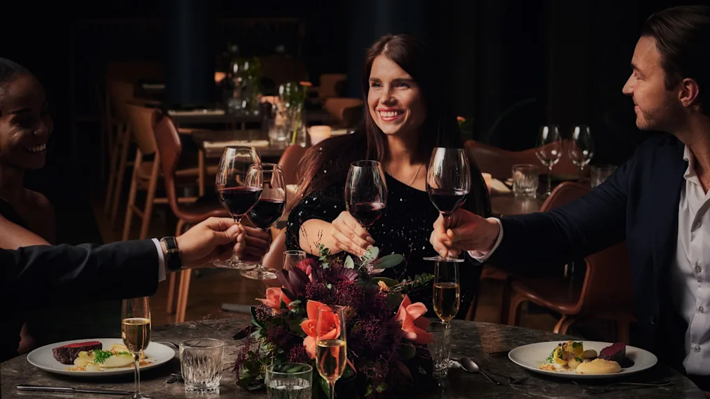 Fire mennesker skåler med vinglass ved et spisebord pyntet med dekorative blomster, omgitt av elegante spisebordsstoler, noe som tyder på en festlig middag i en eksklusiv restaurant.