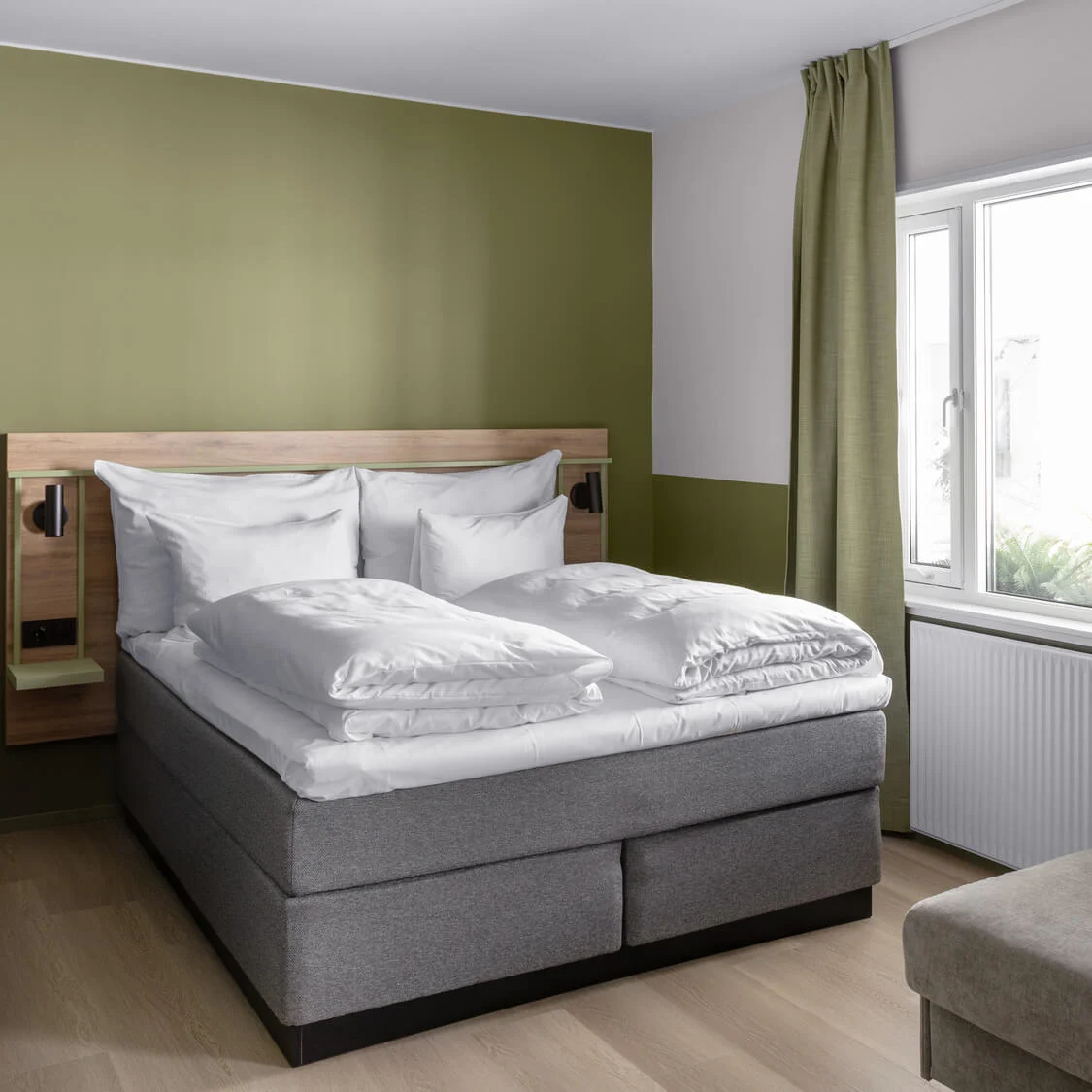En prydligt bäddad dubbelsäng med vita sängkläder står mot en olivgrön vägg, bredvid ett fönster med gröna gardiner, i ett modernt sovrum med en grå soffa.