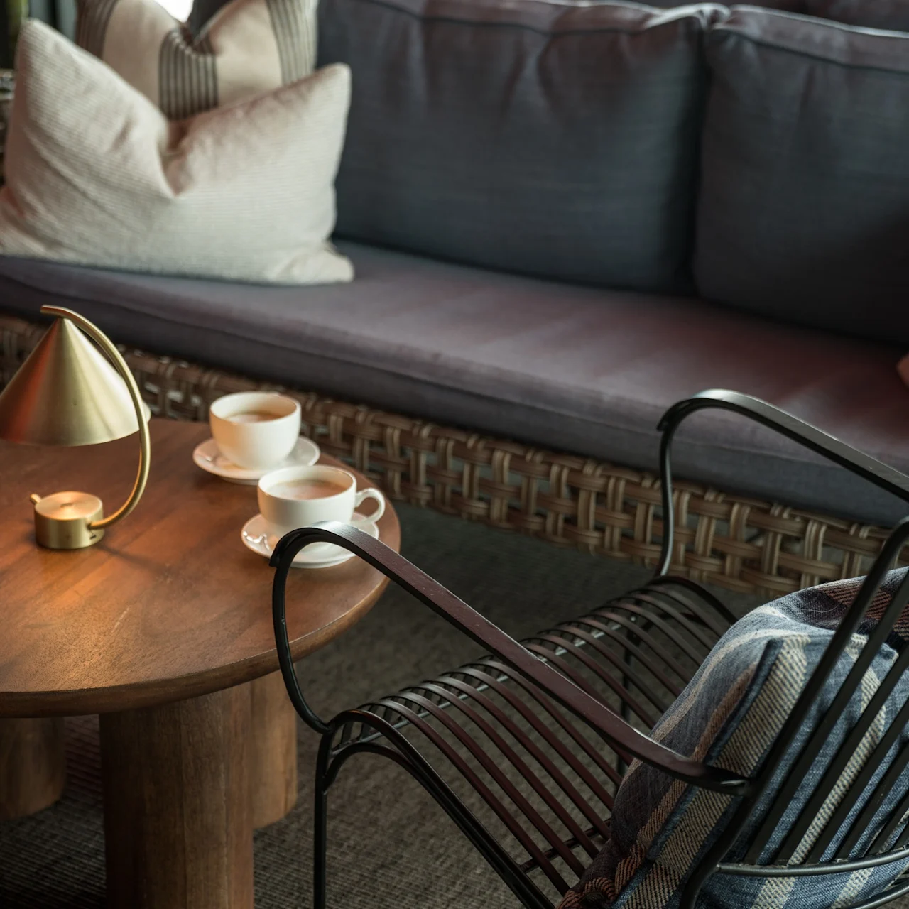 Ett träbord har två kaffekoppar bredvid en liten gyllene lampa, beläget i en mysig sittplats med en vadderad rottingsoffa och metallstol med kudde.