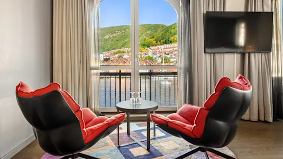 To røde pude stole vender mod et lille bord med to glas, placeret foran et stort vindue med udsigt over farverige bygninger, vand og en grøn bjergside.