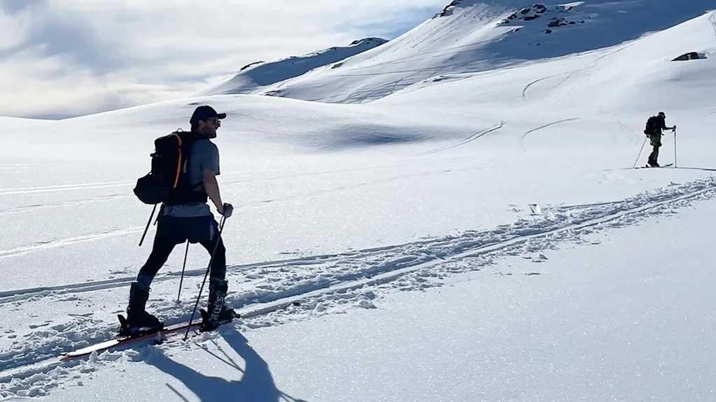 En person står på ski op ad snedækkede bjerge under en klar blå himmel. En anden skiløber er synlig i det fjerne, med skygger på sneen.
