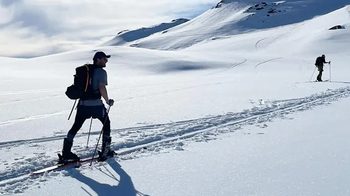 A person skiing uphill in snowy mountains under a clear blue sky. Another skier is visible in the distance, with shadows cast on the snow.