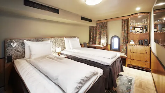To pent oppredde enkeltsenger står side ved side under en taklampe. Et skrivebord og minibar står ved et lite vindu med mønstrede gardiner.