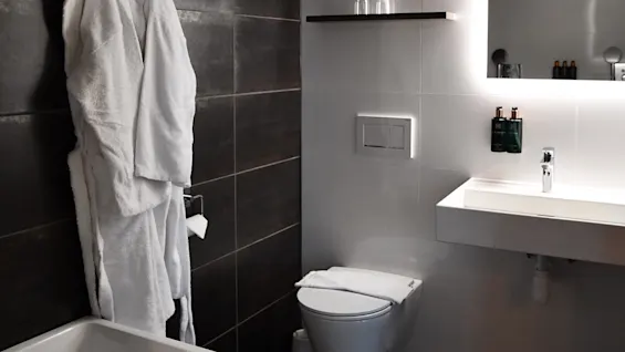 Valkoinen kylpytakki riippuu tummalla laattaseinällä kylpyammeen vieressä modernissa kylpyhuoneessa. Allas ja WC ovat vierekkäin, hyllyillä on hygieniatuotteita ja valaistu peili.