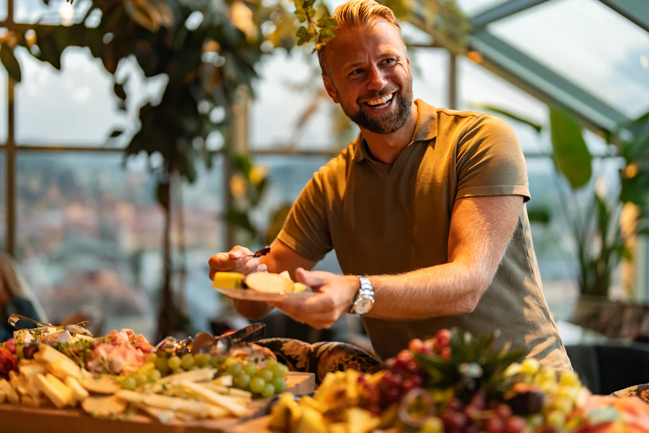 Henkilö ottaa iloisesti ruokaa runsaasta noutopöydästä, jossa on juustoja, hedelmiä ja muita herkullisia välipaloja.