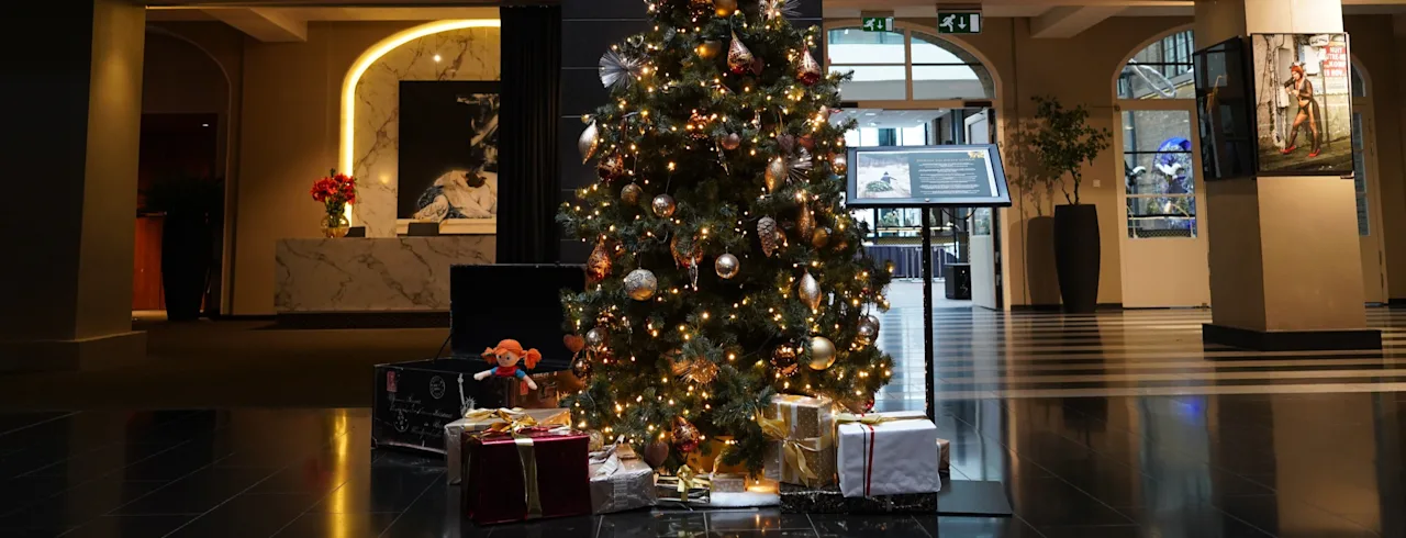 Ett dekorerat julgran står smyckad med ljus och prydnader, omgiven av inslagna gåvor, i en rymlig, elegant lobby med marmordetaljer och blomsterarrangemang.