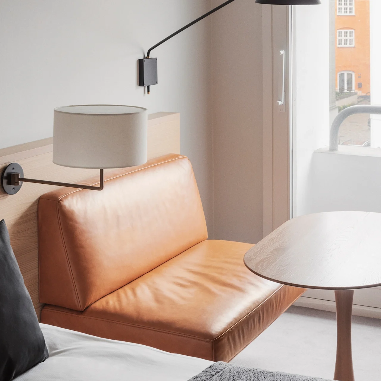 A tan leather sofa sits beside a wooden table under a black wall lamp, near a window overlooking an orange building.