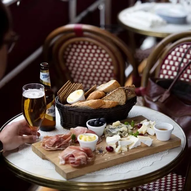 Charcuterie board on a café table features assorted cheeses, olives, cured meats, crackers, and bread. A hand holds a glass of beer beside a bottle labeled "Miller." Wicker chairs surround.