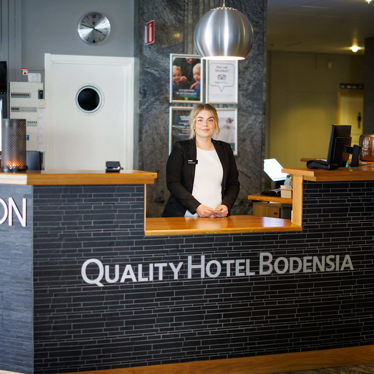 En receptionist står smilande bakom hotellreceptionen i en modern lobby. Skylten på disken lyder: “QUALITY HOTEL BODENSIA.” En väggklocka och reklamaffischer syns i bakgrunden.