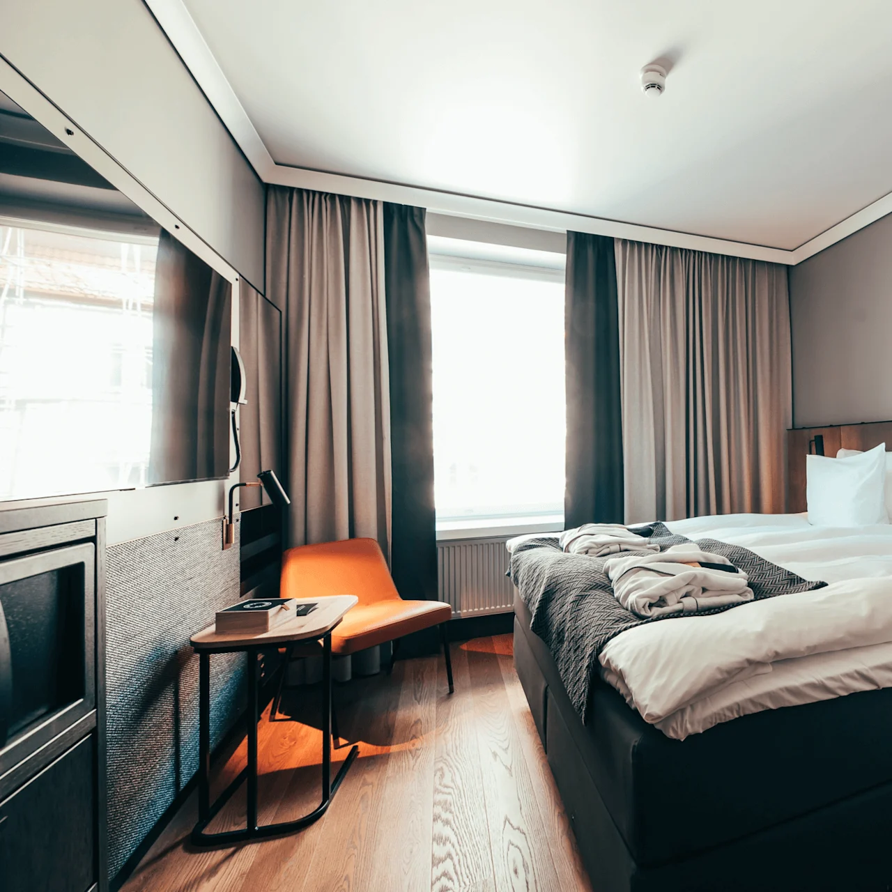Seng med hvitt sengetøy står i et moderne rom, flankert av et lite bord og en oransje stol. Et vindu lyser opp det minimalistiske interiøret, med en veggskulptur over.