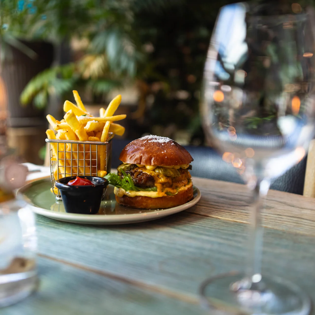 Burger og pommes frites sidder på et træbord i en restaurant, omgivet af et lys og vinglas, med en baggrund af blade.