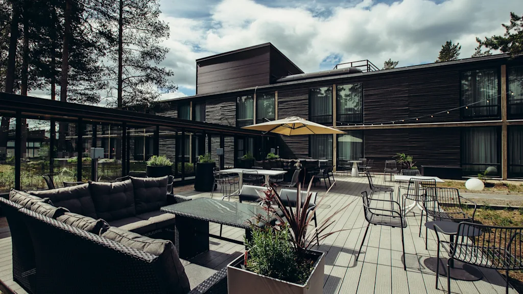 Moderni ulkoterassi, jossa on rottinkisohvia ja lasipöytiä suuren keltaisen aurinkovarjon alla. Tumma puinen rakennus ja korkeat puut ympäröivät aluetta pilvisen taivaan alla.