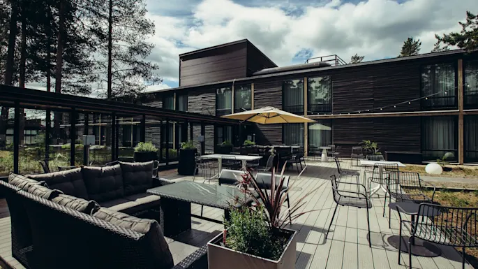Moderni ulkoterassi, jossa on rottinkisohvia ja lasipöytiä suuren keltaisen aurinkovarjon alla. Tumma puinen rakennus ja korkeat puut ympäröivät aluetta pilvisen taivaan alla.