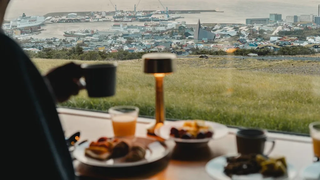 Person holding a cup over a breakfast table, featuring plates with food and a glass of juice. In the background, a seaside town with colorful houses and docked ships.