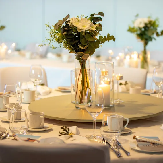 Elegant rundt dekket bord til en festlig anledning med hvite blomster, grønne blader og tente stearinlys.